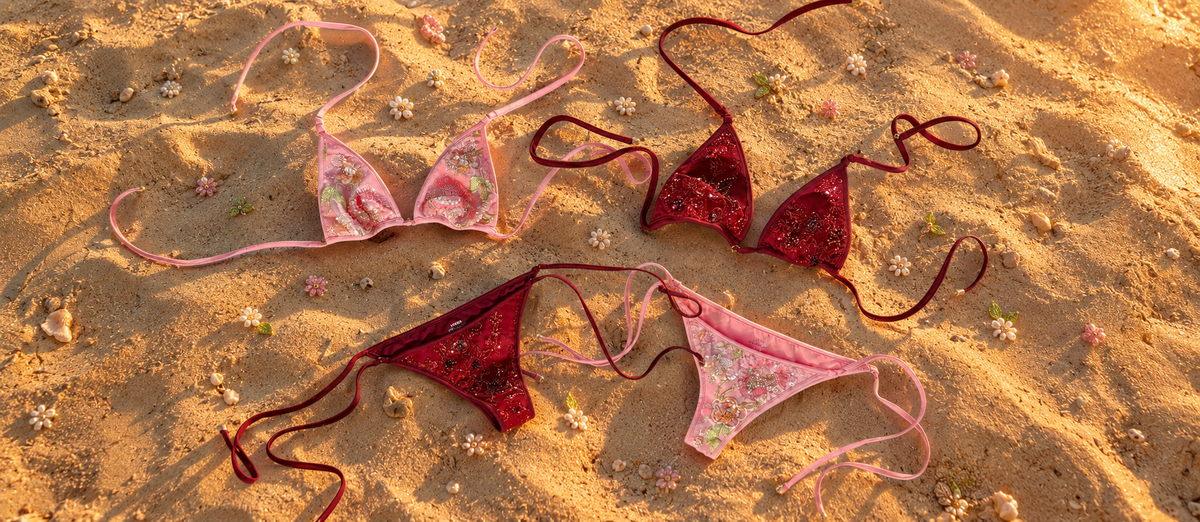 Pink and crimson floral embellished swimwear sets displayed on sand at sunset